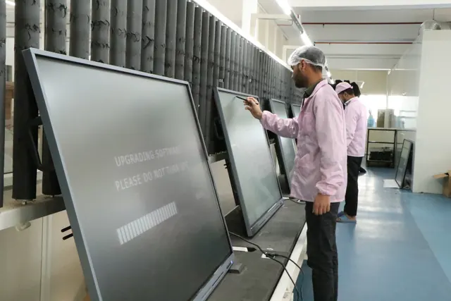 Technicians testing a row of interactive panels on the factory floor, the nearest screen showing a software-upgrade message