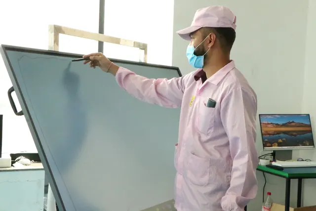 Technician in a factory coat, cap and face mask checking an interactive panel's screen with a pen